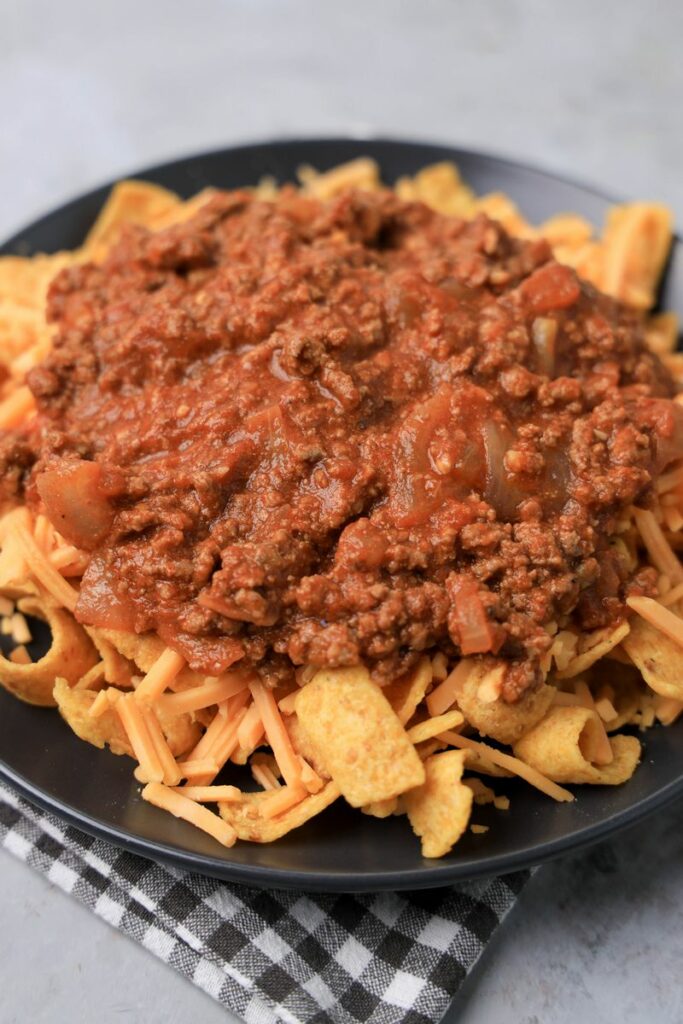 Frito pie on a gray plate on a gray plaid napkin on a faux concrete backdrop.