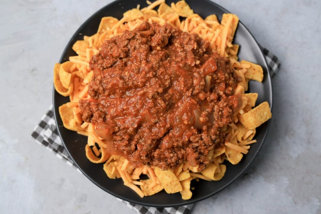 Frito pie on a gray plate on a gray plaid napkin on a faux concrete backdrop.