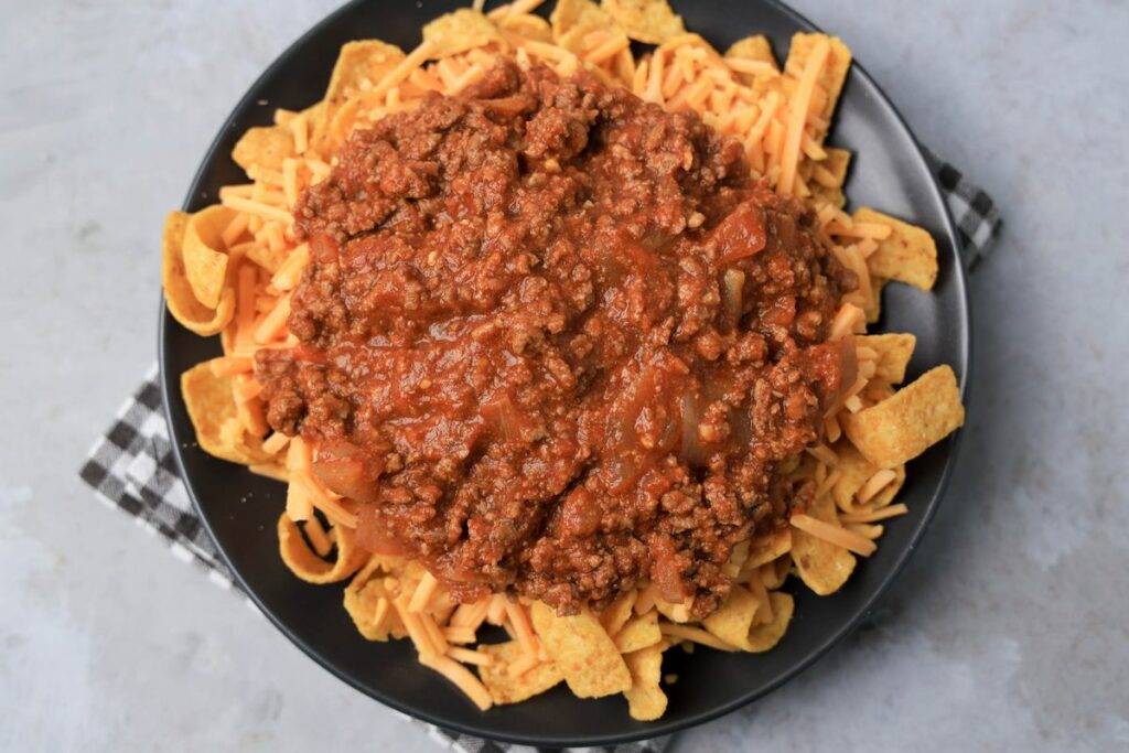 Frito pie on a gray plate on a gray plaid napkin on a faux concrete backdrop.