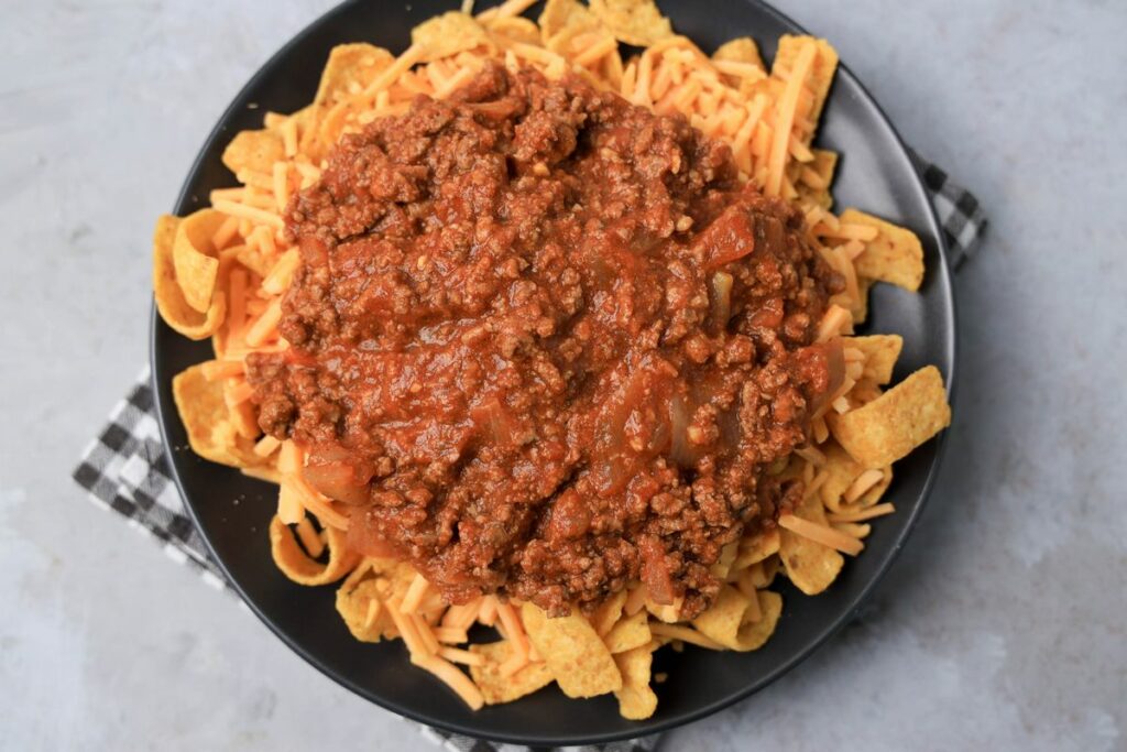 Frito pie on a gray plate on a gray plaid napkin on a faux concrete backdrop.