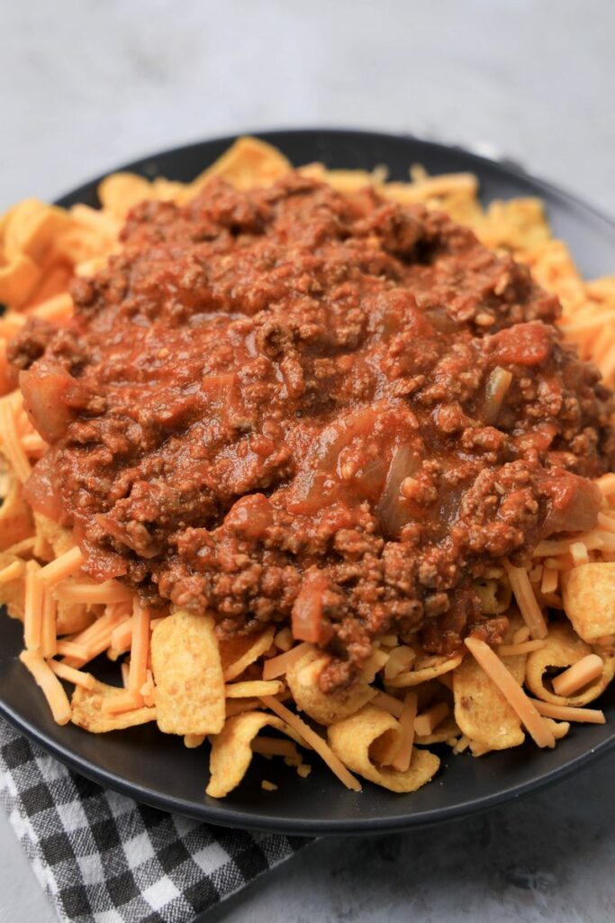 Frito pie on a gray plate on a gray plaid napkin on a faux concrete backdrop.