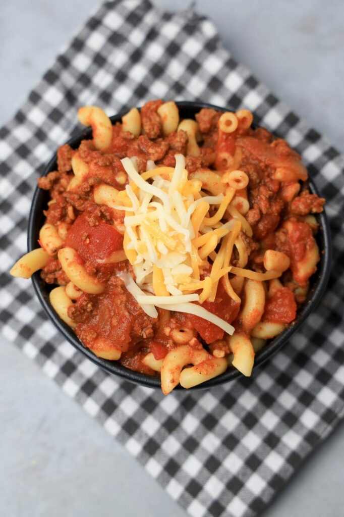 Chili mac with elbow pasta and round beef in a black bowl topped with cheese. On a plaid napkin.