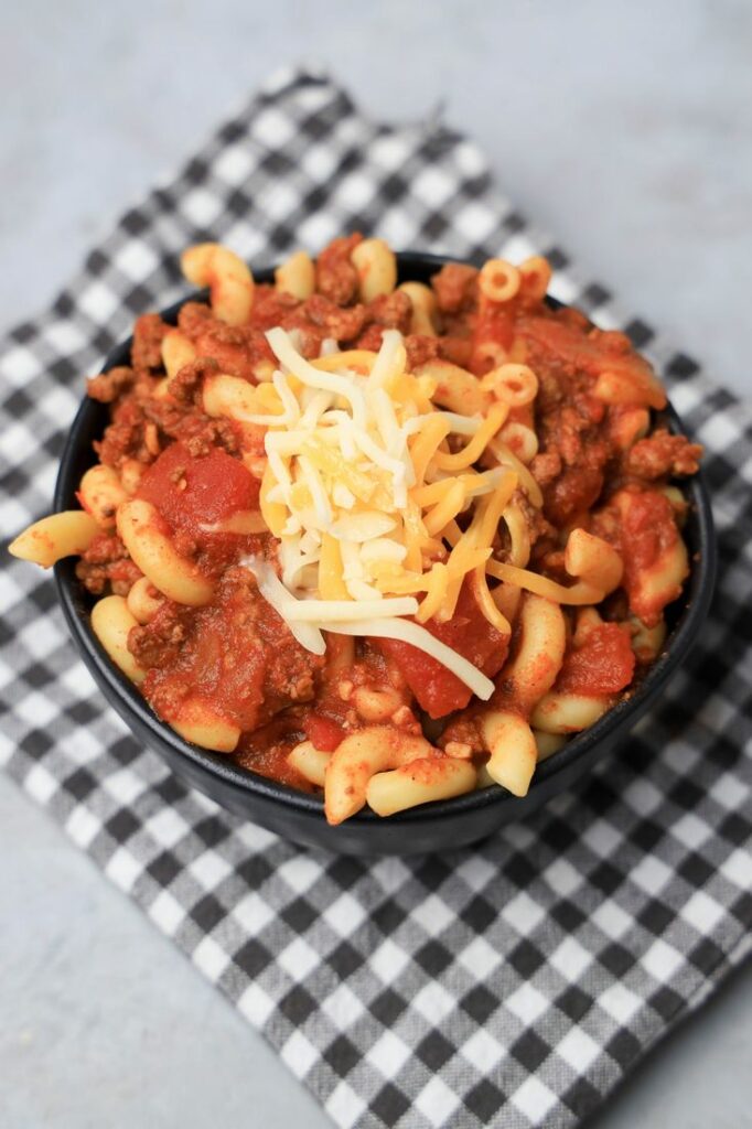 Chili mac with elbow pasta and round beef in a black bowl topped with cheese. On a plaid napkin.
