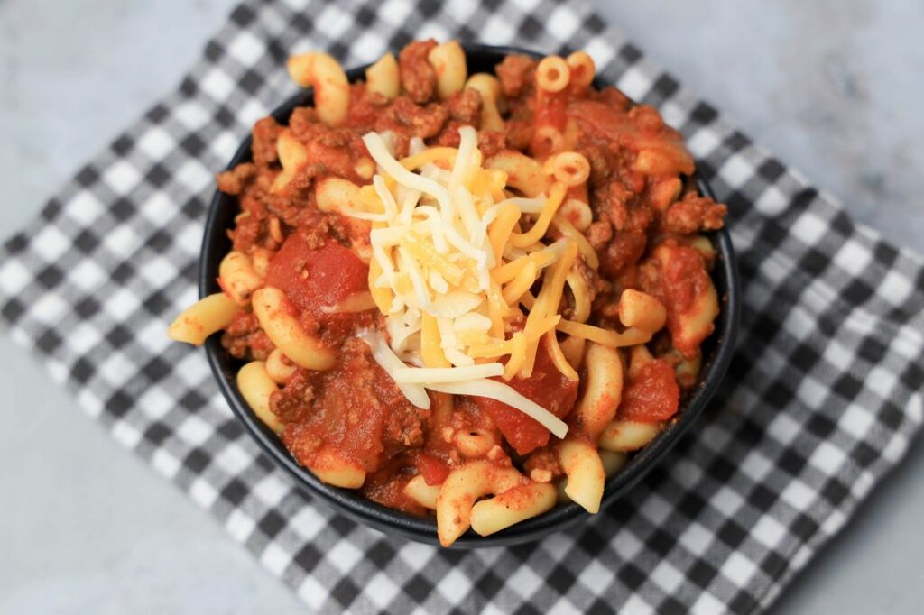 Chili mac with elbow pasta and round beef in a black bowl topped with cheese. On a plaid napkin.