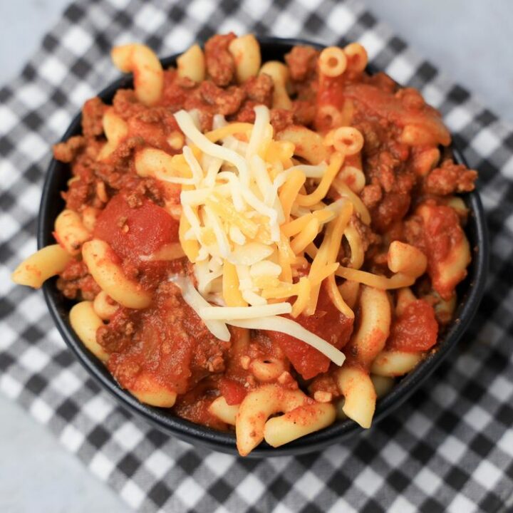 Chili mac with elbow pasta and round beef in a black bowl topped with cheese. On a plaid napkin.