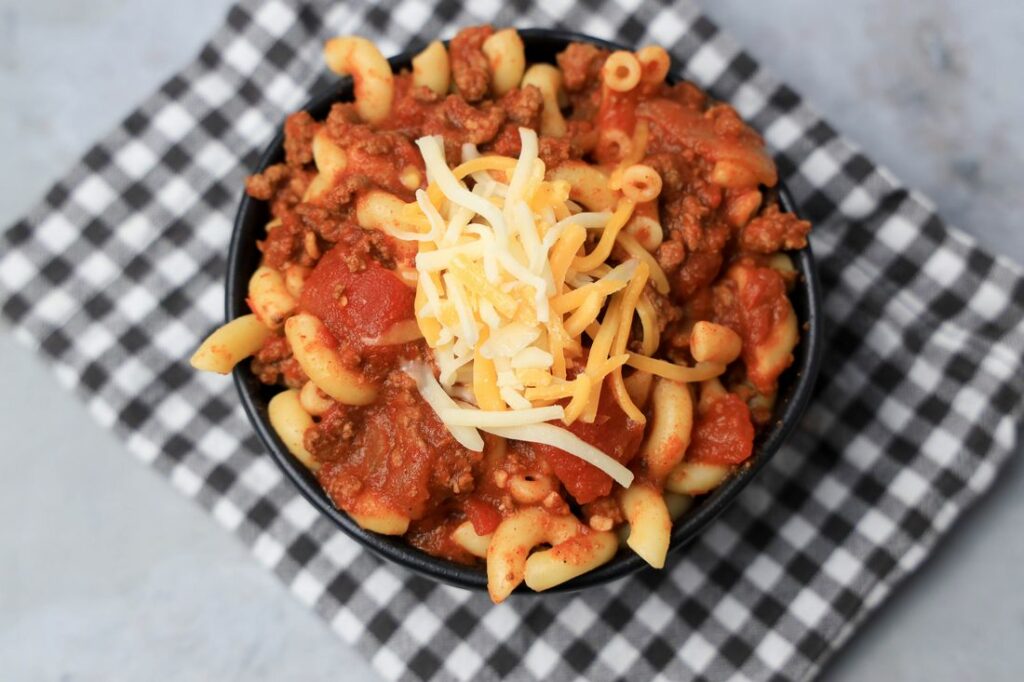 Chili mac with elbow pasta and round beef in a black bowl topped with cheese. On a plaid napkin.