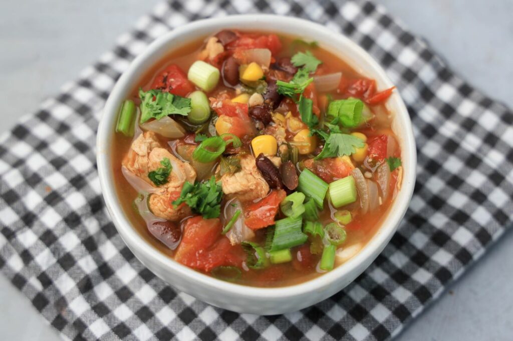 Baja Chicken Soup in a white bowl on a gray plaid napkin on a faux concrete backdrop.