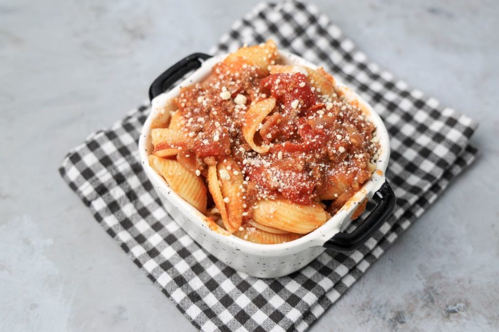 Italian Goulash with stewed tomatoes, onions, and bell pepper topped with parmesan cheese in a white bowl on a gray plaid napkin and concrete backdrop