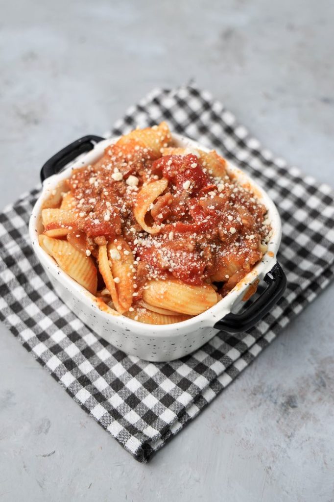 Italian Goulash with stewed tomatoes, onions, and bell pepper topped with parmesan cheese in a white bowl on a gray plaid napkin and concrete backdrop