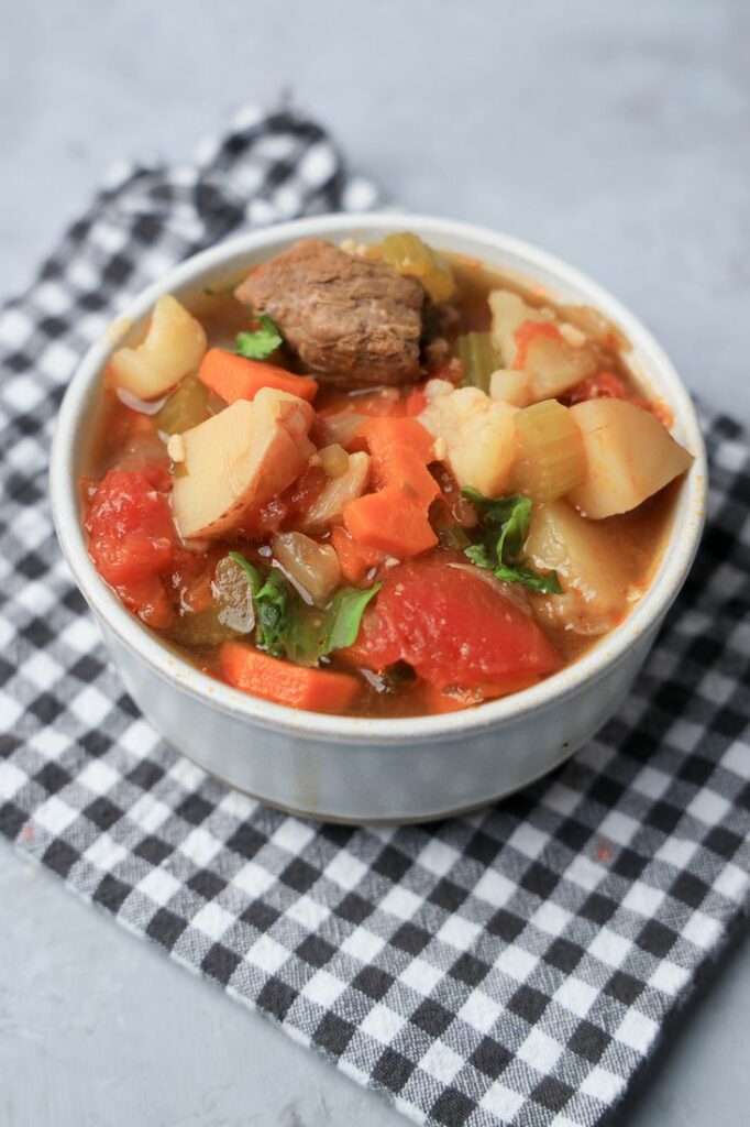 Instant pot tuscan beef stew in a white bowl on a gray plaid napkin on a faux concrete back drop.