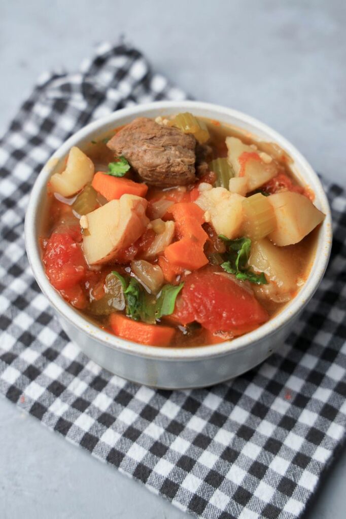 Instant pot tuscan beef stew in a white bowl on a gray plaid napkin on a faux concrete back drop.