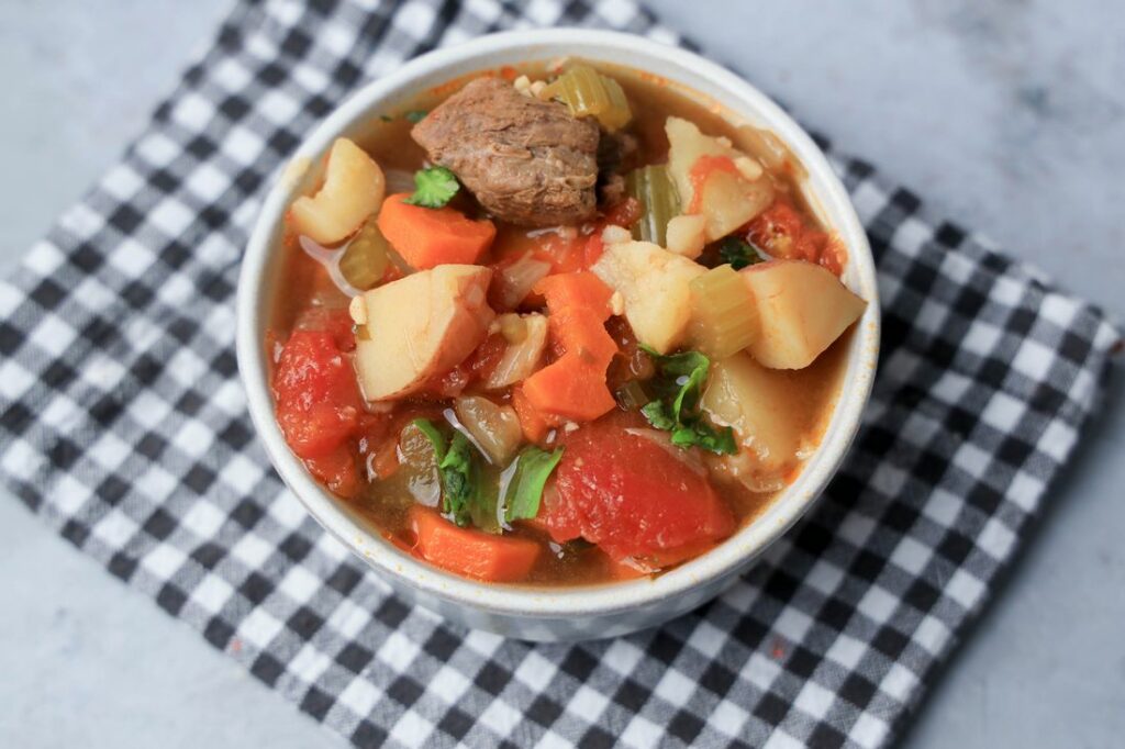 Instant pot tuscan beef stew in a white bowl on a gray plaid napkin on a faux concrete back drop.