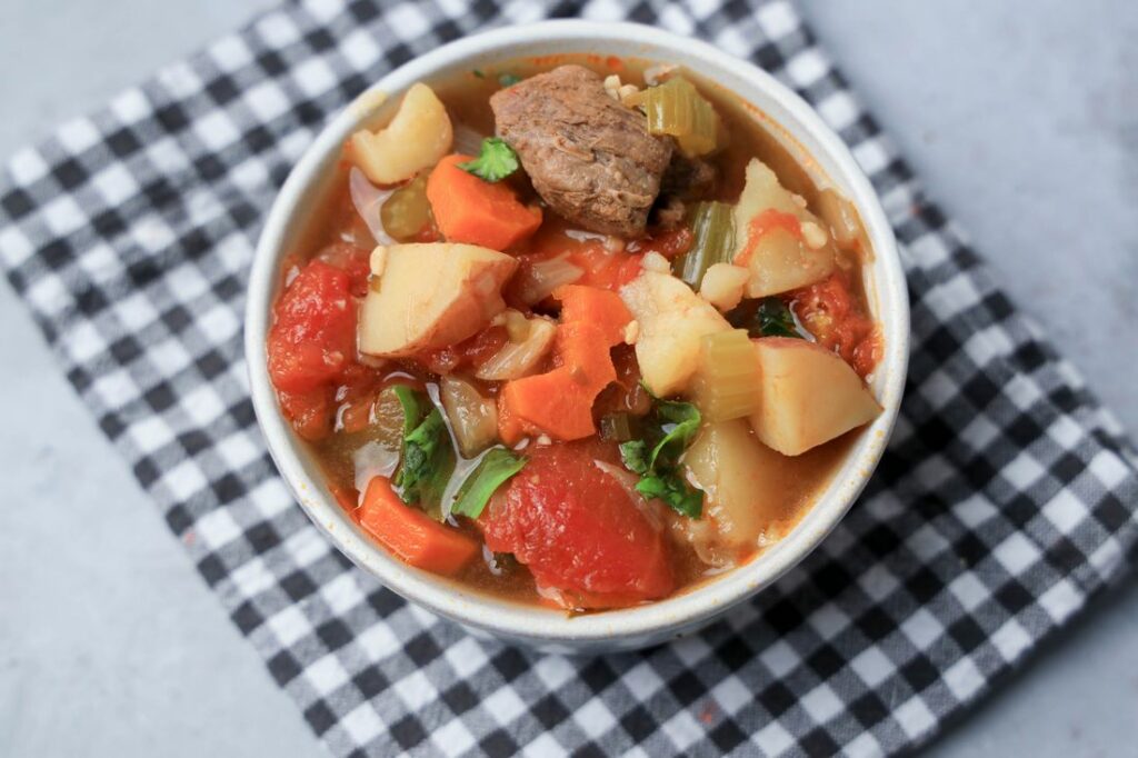 Instant pot tuscan beef stew in a white bowl on a gray plaid napkin on a faux concrete back drop.