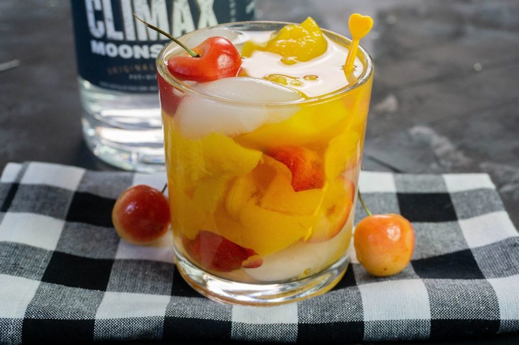 Cocktail with rainier cherries on black plaid napkin with moonshine bottle in the back