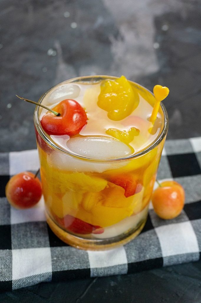 Rainier Cherry Peach Moonshine Cocktail on gray background with rainier cherries and a plaid napkin