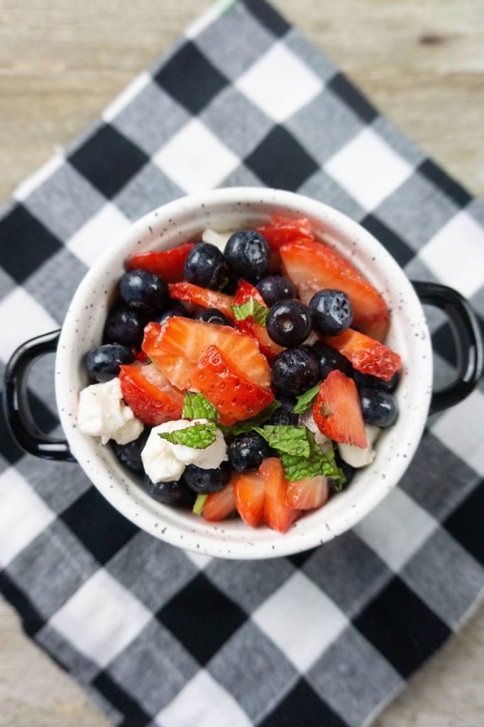 July 4th Fruit Salad with strawberries, blueberries, and mozzarella pearl cheese in a whote bowl with black speckles on a black and white plaid napkin