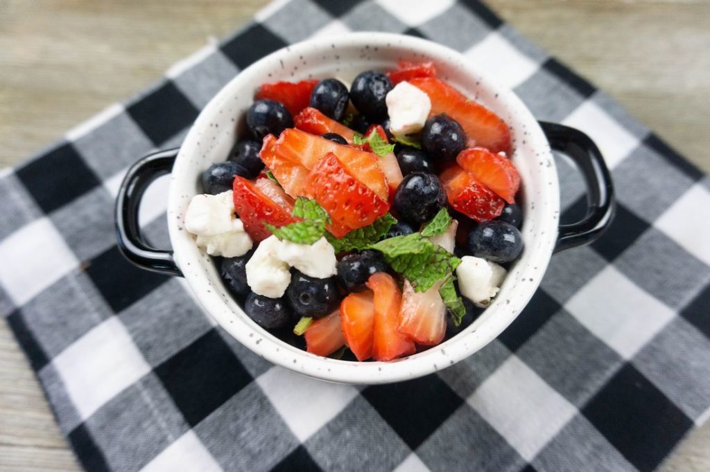 July 4th Fruit Salad with strawberries, blueberries, and mozzarella pearl cheese in a whote bowl with black speckles on a black and white plaid napkin