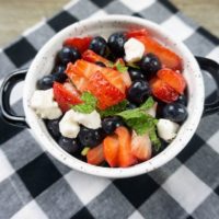 July 4th Fruit Salad with strawberries, blueberries, and mozzarella pearl cheese in a whote bowl with black speckles on a black and white plaid napkin