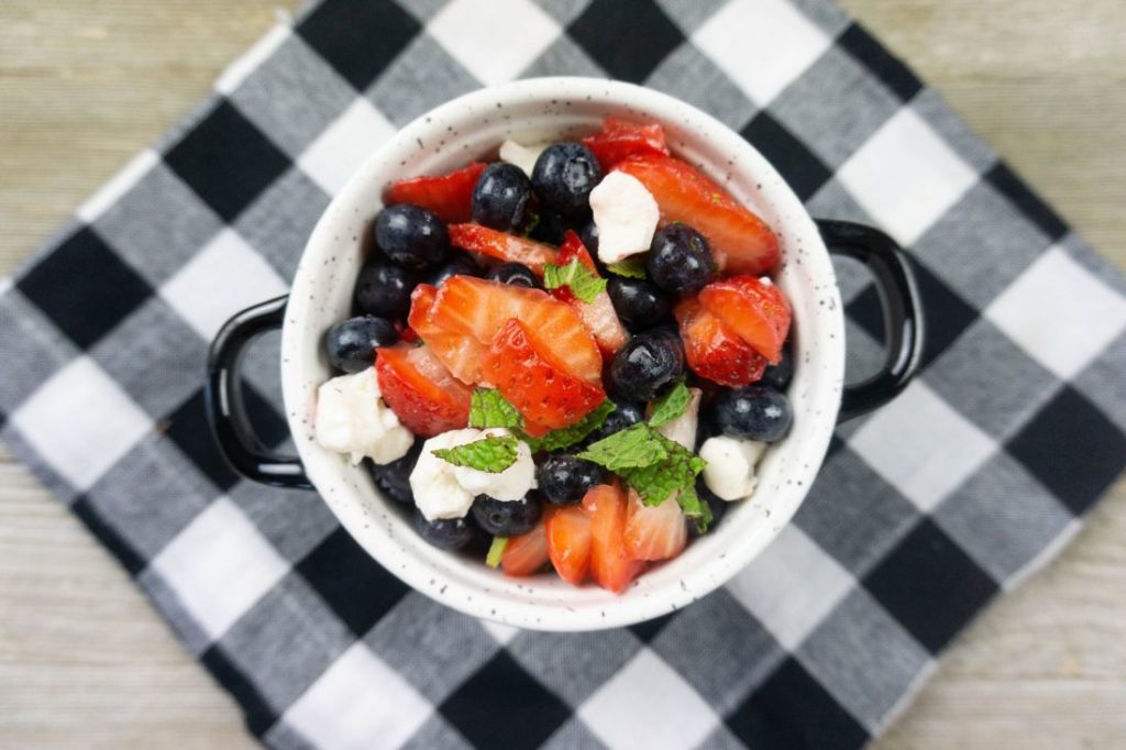 July 4th Fruit Salad with strawberries, blueberries, and mozzarella pearl cheese in a whote bowl with black speckles on a black and white plaid napkin