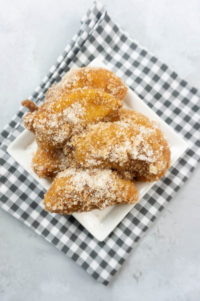 Deep fried apples on a white plate with cinnamon sugar on top on a plaid napkin and concrete backdrop