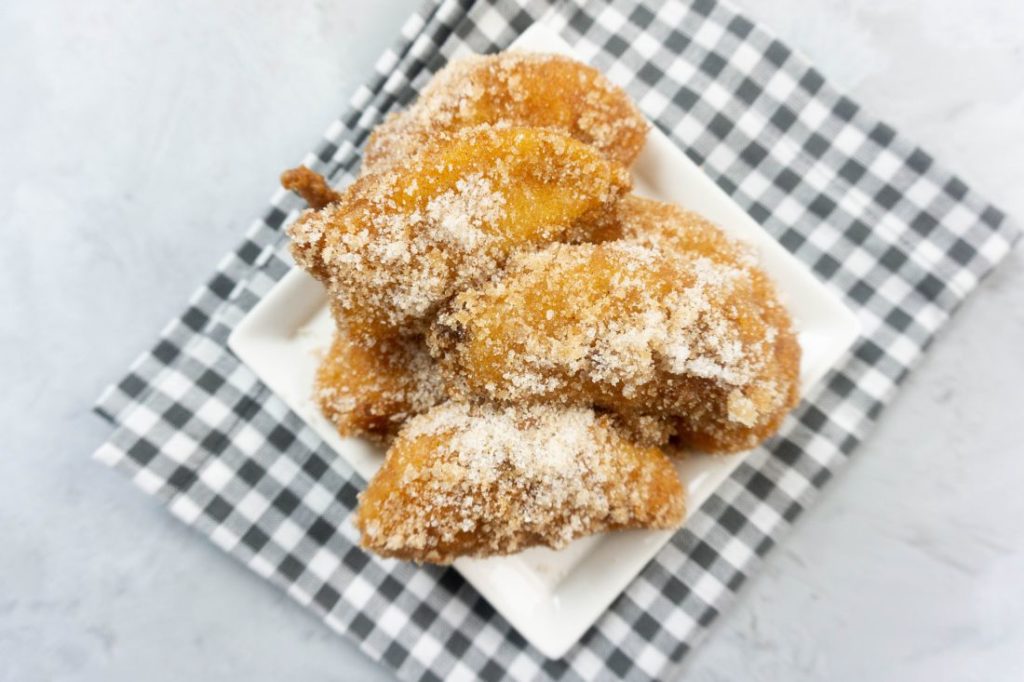 Deep fried apples on a white plate with cinnamon sugar on top on a plaid napkin and concrete backdrop