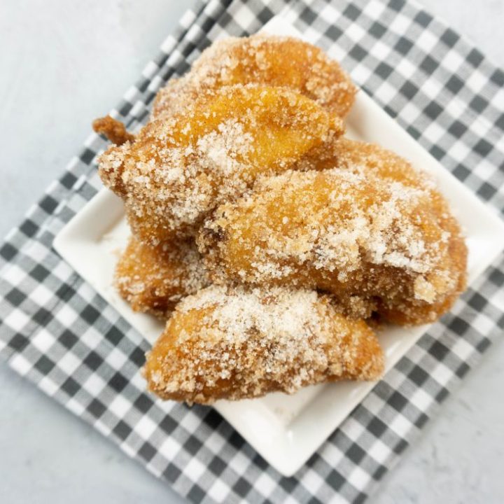 Deep fried apples on a white plate with cinnamon sugar on top on a plaid napkin and concrete backdrop