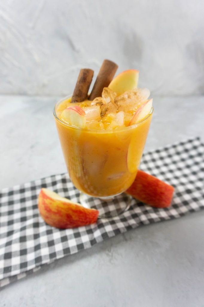 Cloudy Pumpkin Patch Cocktail with apples and pumpkin topped with cinnamon sticks in a glass on gray plaid napkin on a concrete backdrop