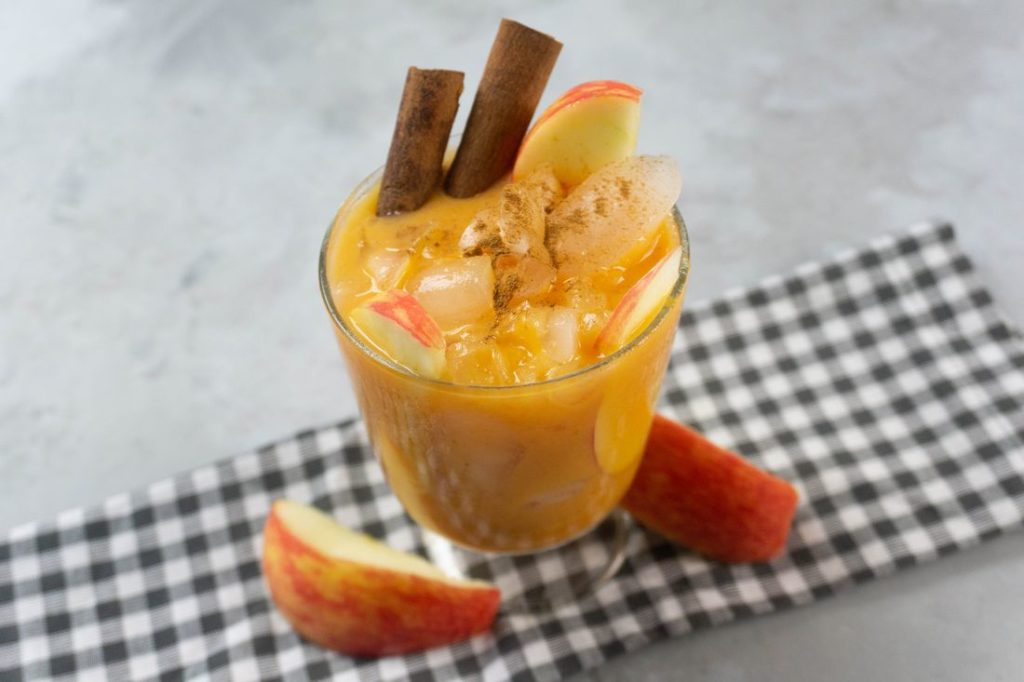 Cloudy Pumpkin Patch Cocktail with apples and pumpkin topped with cinnamon sticks in a glass on gray plaid napkin on a concrete backdrop