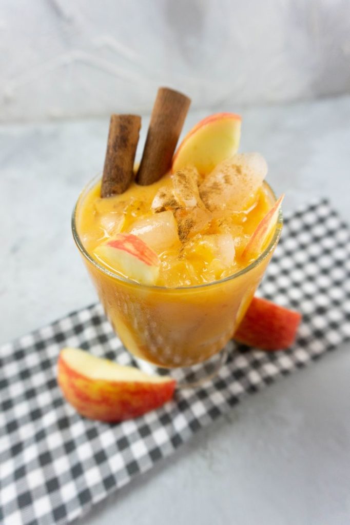 Cloudy Pumpkin Patch Cocktail with apples and pumpkin topped with cinnamon sticks in a glass on gray plaid napkin on a concrete backdrop