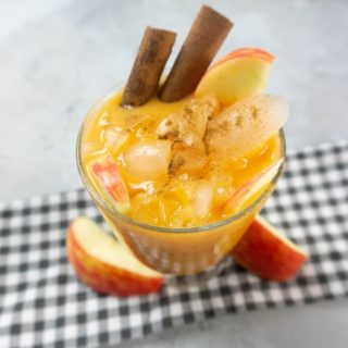 Cloudy Pumpkin Patch Cocktail with apples and pumpkin topped with cinnamon sticks in a glass on gray plaid napkin on a concrete backdrop