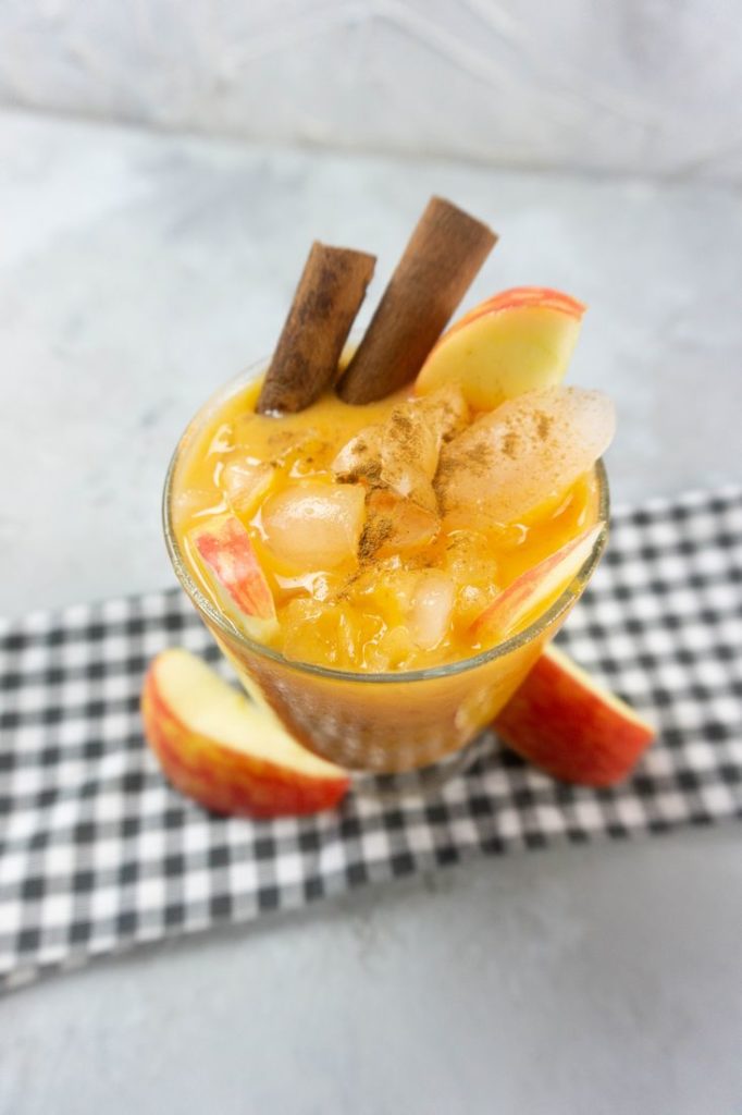 Cloudy Pumpkin Patch Cocktail with apples and pumpkin topped with cinnamon sticks in a glass on gray plaid napkin on a concrete backdrop