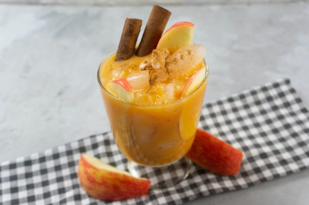 Cloudy Pumpkin Patch Cocktail with apples and pumpkin topped with cinnamon sticks in a glass on gray plaid napkin on a concrete backdrop