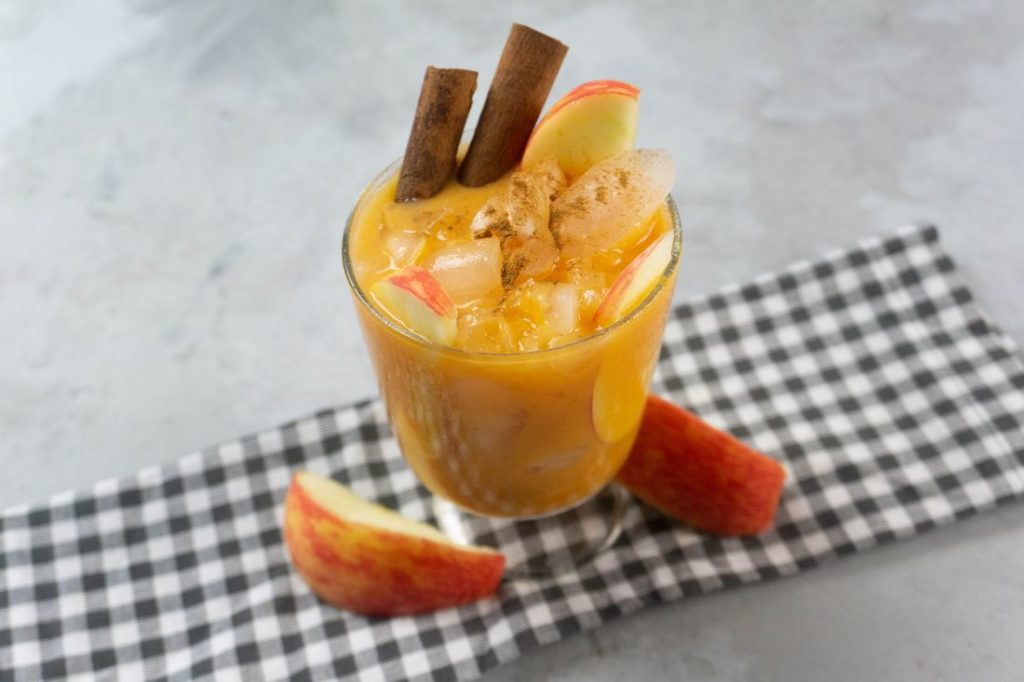 Cloudy Pumpkin Patch Cocktail with apples and pumpkin topped with cinnamon sticks in a glass on gray plaid napkin on a concrete backdrop