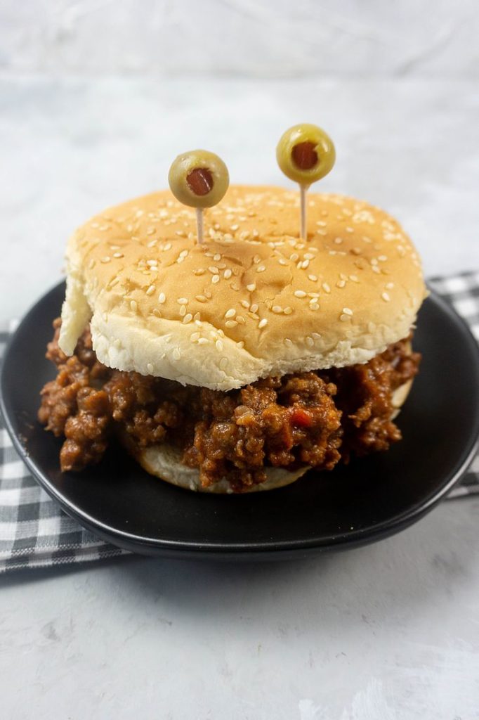 Monster sloppy joe with olive eyes and on a black plate with gray plaid napkin.