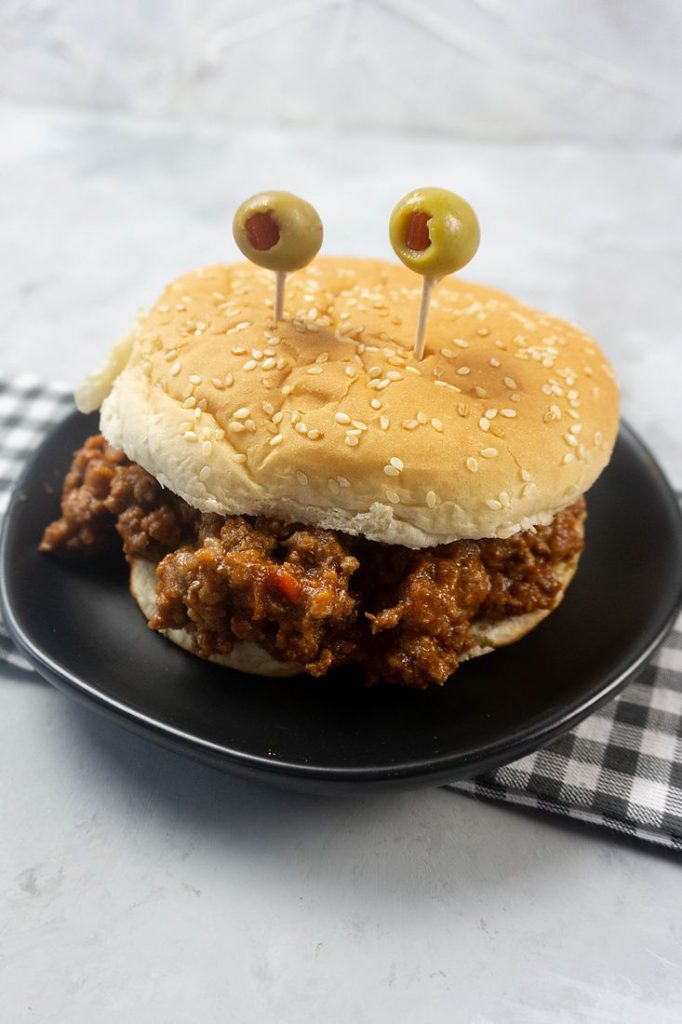 Monster sloppy joe with olive eyes and on a black plate with gray plaid napkin.