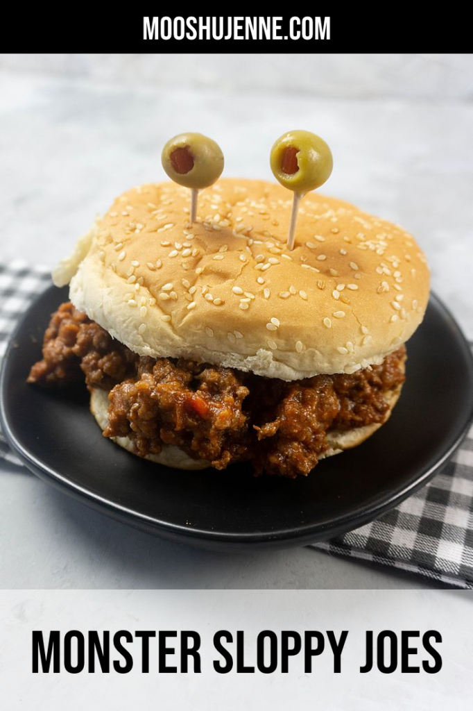 Sloppy joe on a bun with olives on toothpicks for eyes on a gray plaid napkin and concrete backdrop