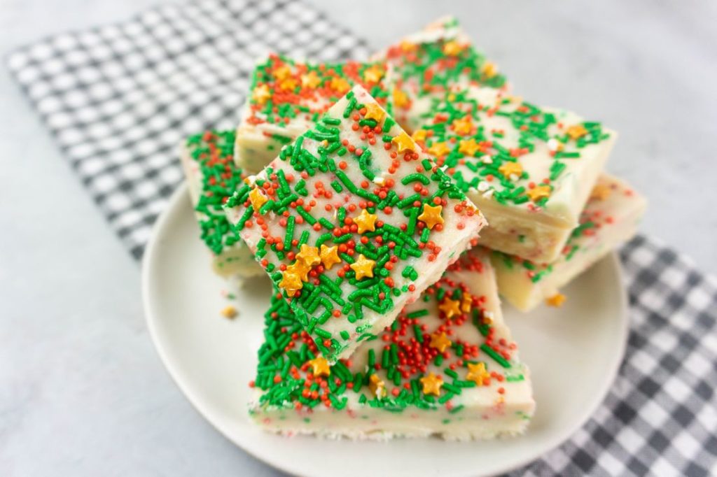 Sugar cookie fudge covered in Christmas sprinkles on a white plate with grey plaid napkin on a concrete back drop