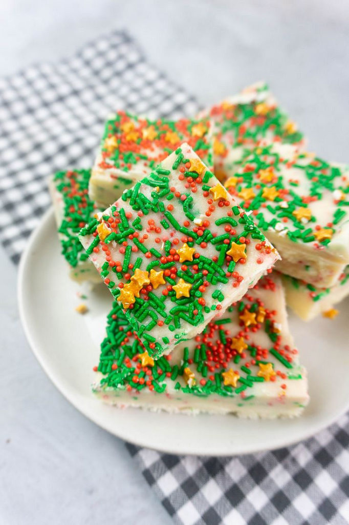 Sugar cookie fudge covered in Christmas sprinkles on a white plate with grey plaid napkin on a concrete back drop