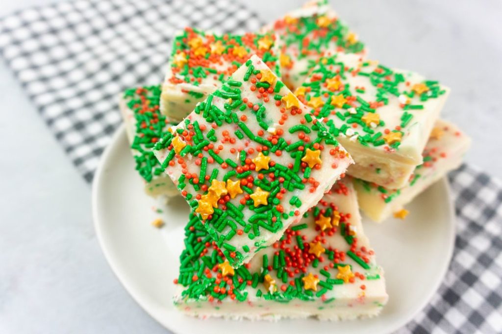 Sugar cookie fudge covered in Christmas sprinkles on a white plate with grey plaid napkin on a concrete back drop