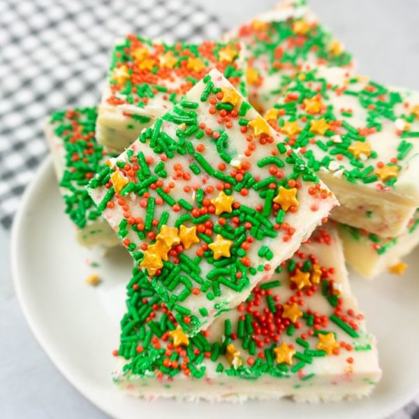 Sugar cookie fudge covered in Christmas sprinkles on a white plate with grey plaid napkin on a concrete back drop