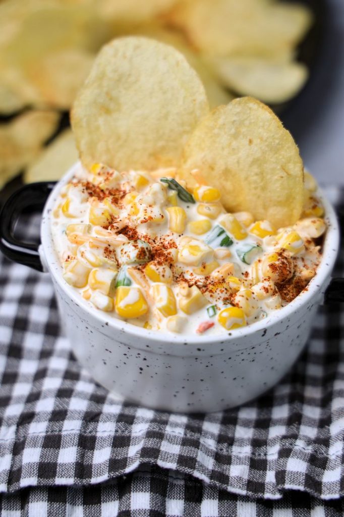 Southwestern Corn Dip in a white and black speckled bowl with a gray plaid napkin on a concrete backdrop