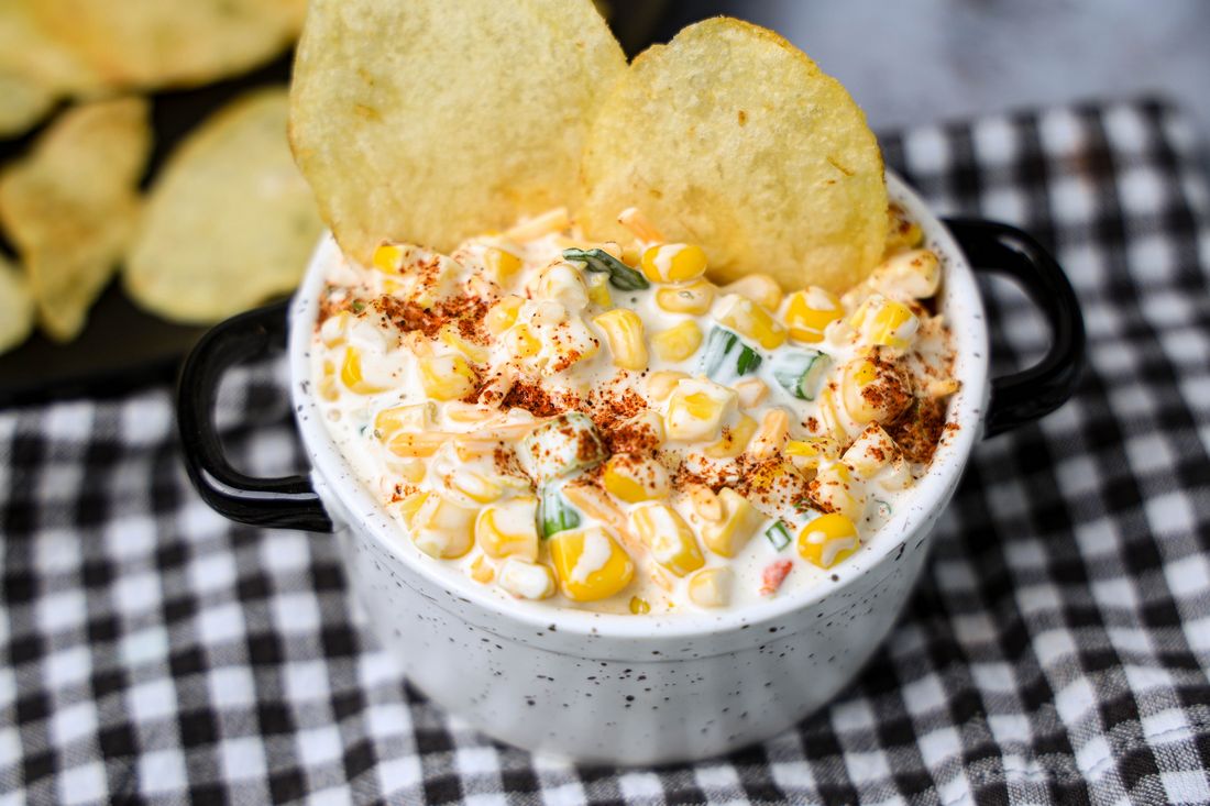 Southwestern Corn Dip in a white and black speckled bowl with a gray plaid napkin on a concrete backdrop