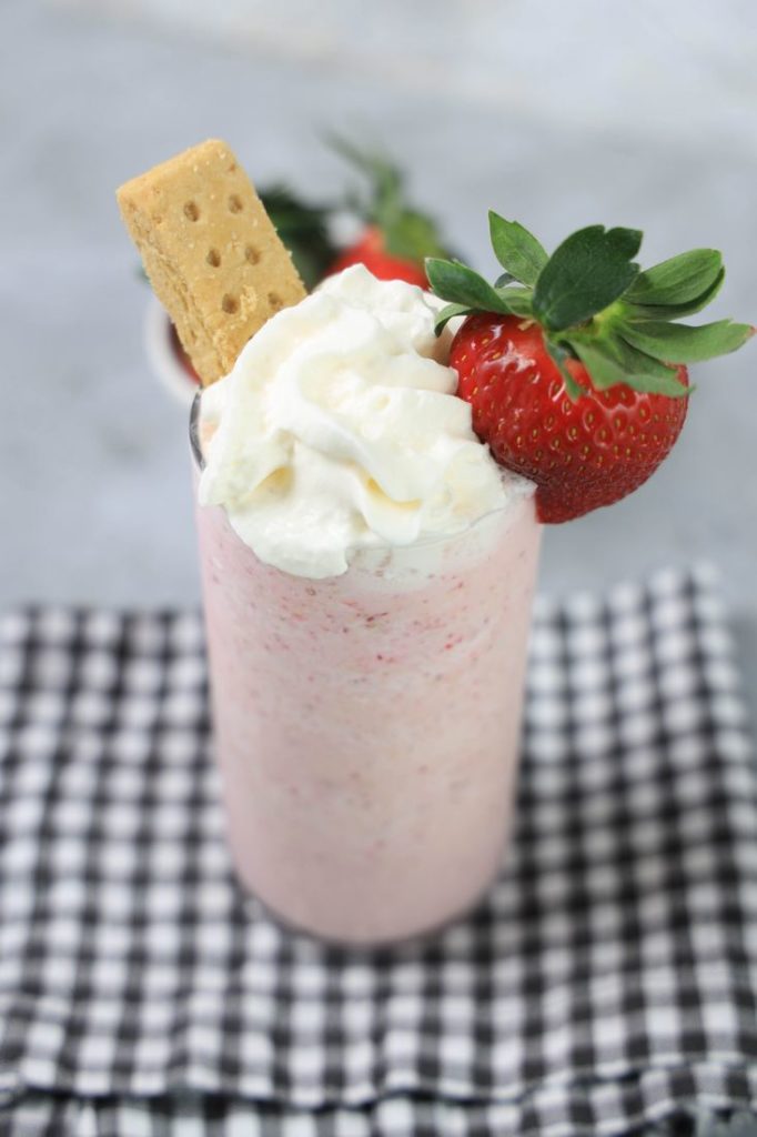 Strawberry Shortcake Shake in a glass with strawberry on top on a gray plaid napkin on a concrete backdrop