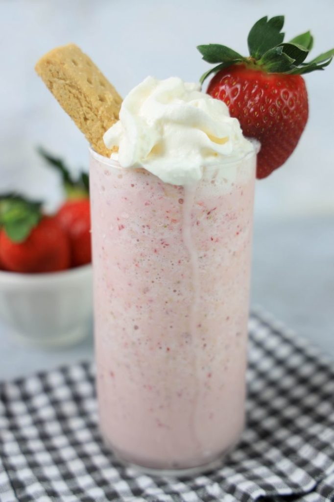 Strawberry Shortcake Shake in a glass with strawberry on top on a gray plaid napkin on a concrete backdrop