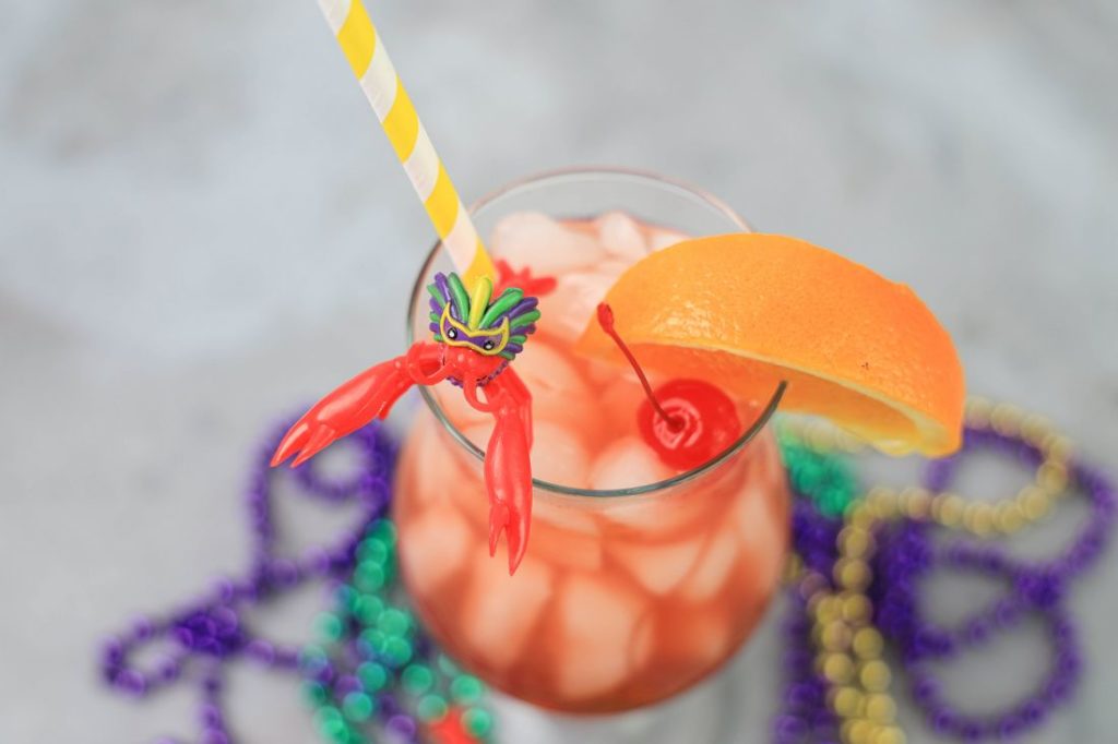 Hurricane cocktail in a hurricane glass with a yellow and white striped straw on a concrete backdrop with mardi gras beads and topped with a cherry, orange slice, and plastic crawfish