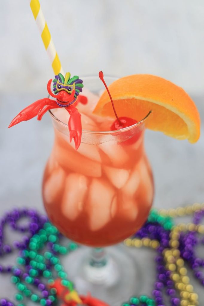 Hurricane cocktail in a hurricane glass with a yellow and white striped straw on a concrete backdrop with mardi gras beads and topped with a cherry, orange slice, and plastic crawfish