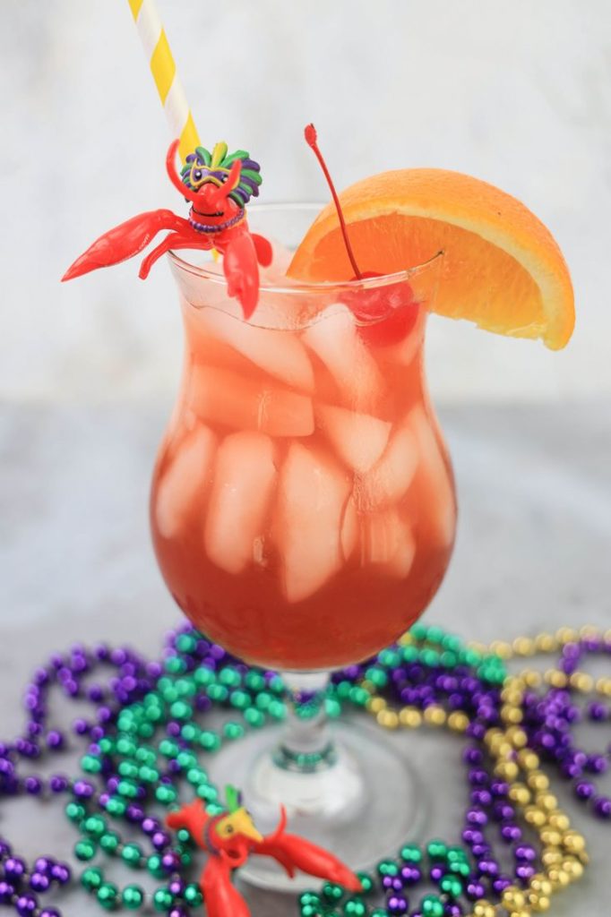 Hurricane cocktail in a hurricane glass with a yellow and white striped straw on a concrete backdrop with mardi gras beads and topped with a cherry, orange slice, and plastic crawfish