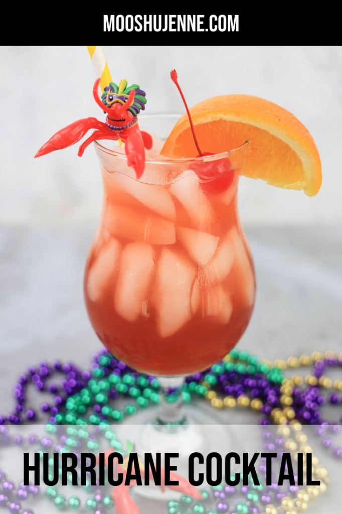 Hurricane cocktail in a hurricane glass with a yellow and white striped straw on a concrete backdrop with mardi gras beads and topped with a cherry, orange slice, and plastic crawfish