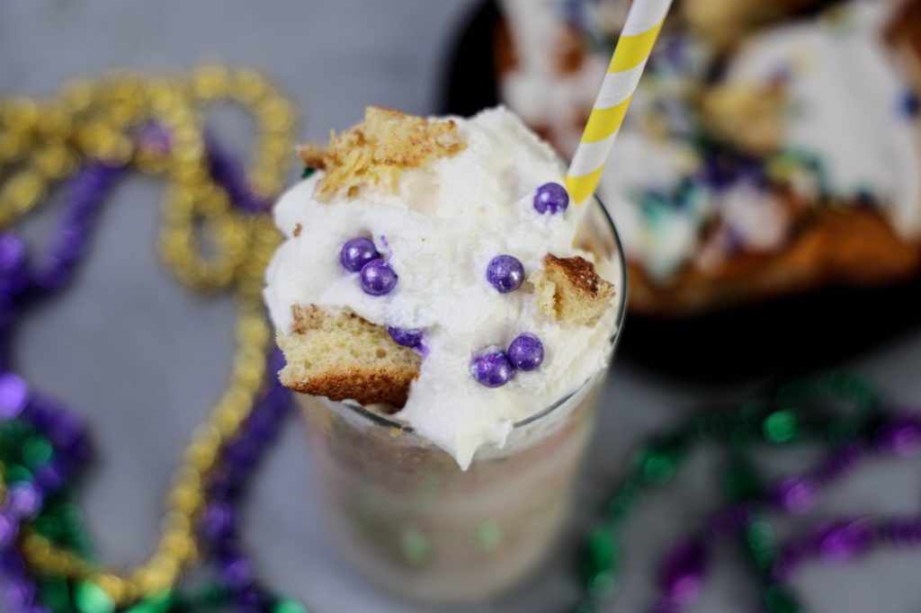 King Cake Frappuccino in a tall glass topped with whipped topping, purple pearl candies, and a yellow straw.