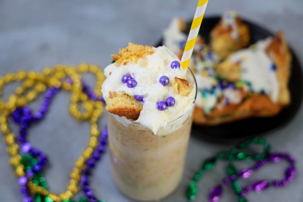 King Cake Frappuccino in a tall glass topped with whipped topping, purple pearl candies, and a yellow straw.